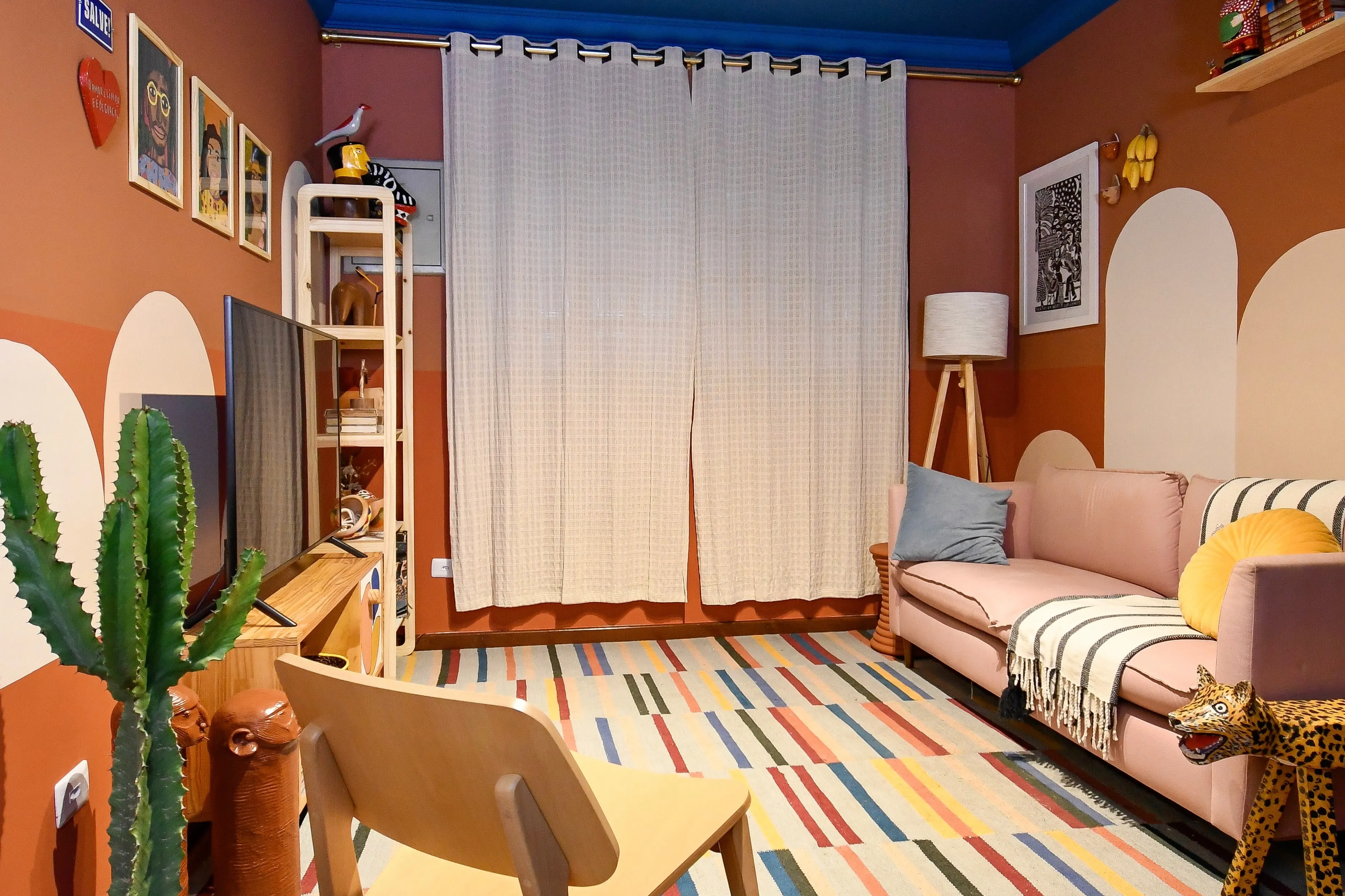 Sala projetada pela Orí: paredes em terracota com arcos pintados, teto azul, sofá rosa, tapete listrado colorido e estante com arte popular brasileira