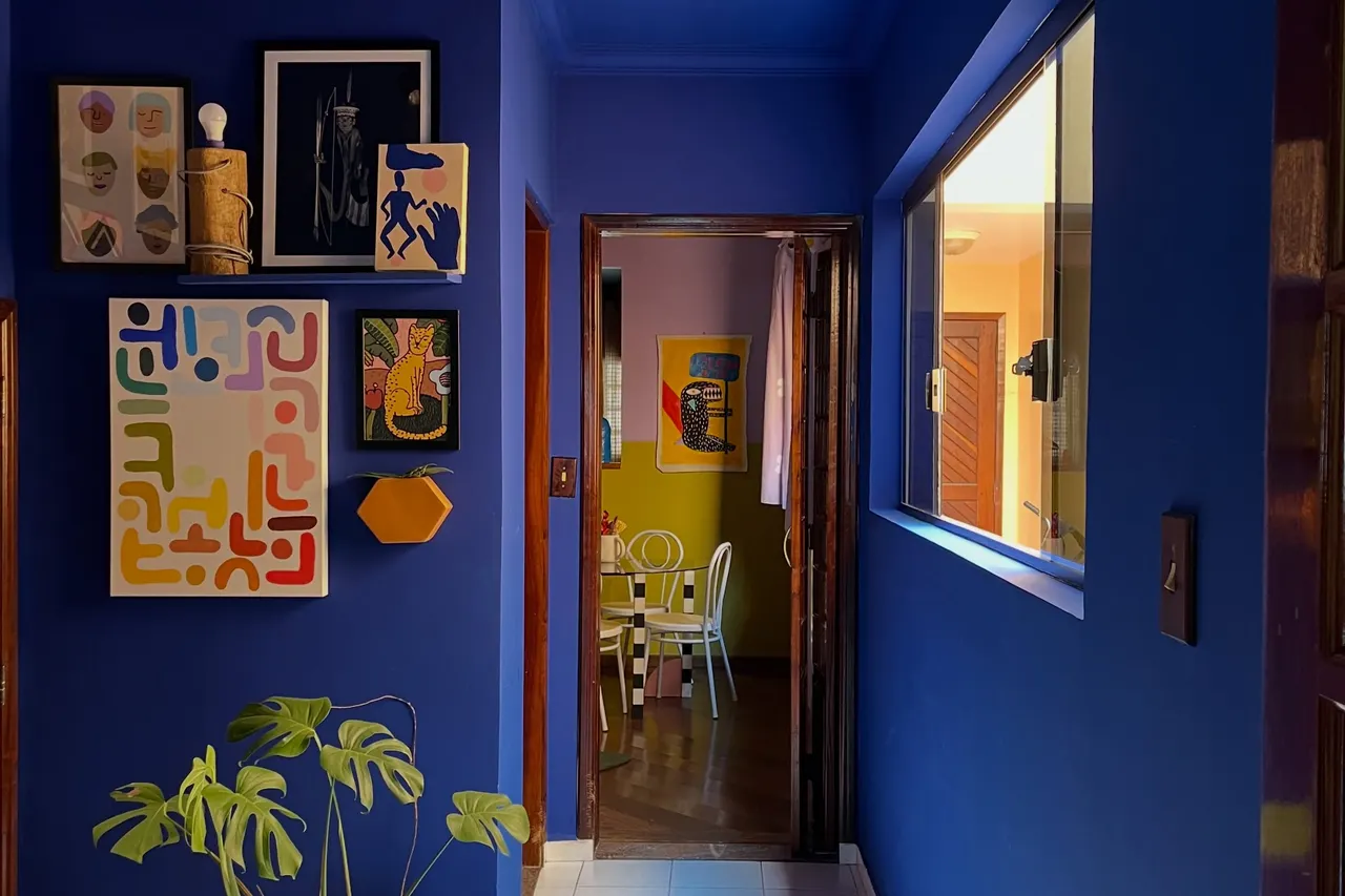 Corredor com paredes azul-cobalto, quadros coloridos e vista para a sala de jantar amarela