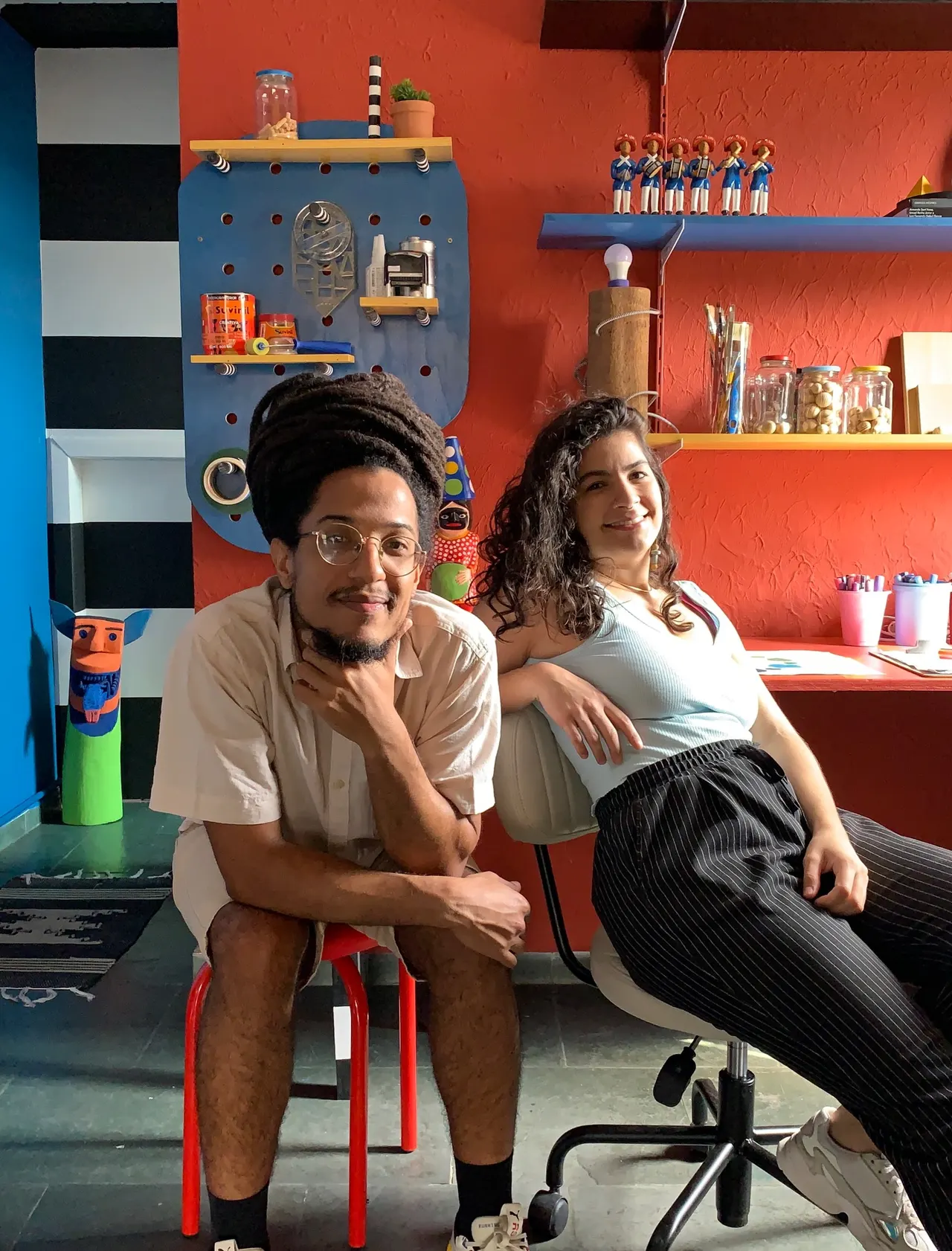 Jorge Felipe e Thaís Melo sentados lado a lado, com parede vermelha e azul, listras pretas e brancas e um pegboard azul ao fundo