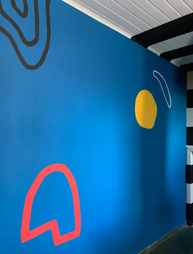 Detalhe de mural em parede azul, com círculo amarelo e arco vermelho pintados à mão