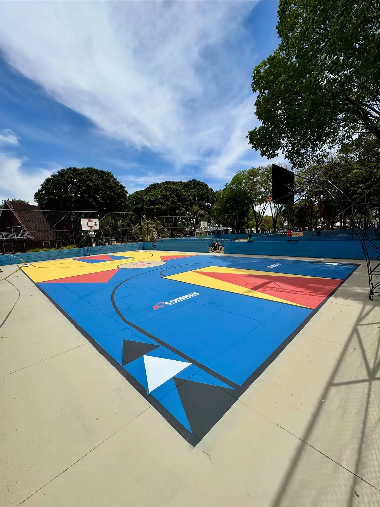 Quadra poliesportiva pública em Minas Gerais, pintada em azul, amarelo e vermelho, com triângulos pretos e brancos e céu azul ao fundo