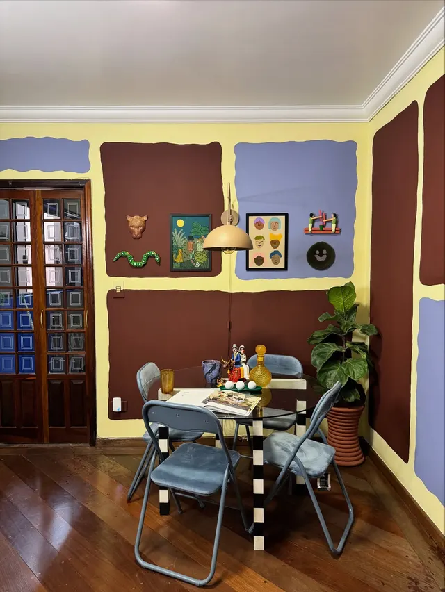 Sala de jantar com parede em blocos vinho, lilás e creme desenhados à mão e mesa com cadeiras azuis