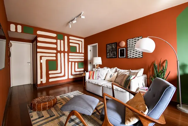 Sala com parede terracota e mural gráfico em faixas brancas e quadrados verdes, sofá claro e poltrona de madeira